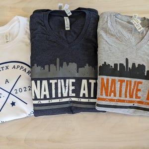 May include: Three V-neck t-shirts featuring a city skyline graphic and the text "NATIVE ATX APPAREL". The shirts are in white, dark grey, and light grey.