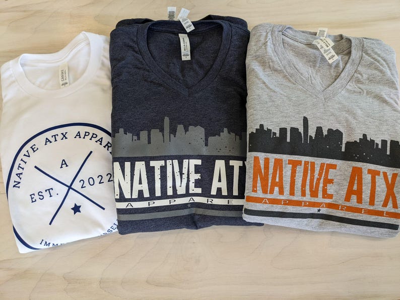 May include: Three V-neck t-shirts featuring a city skyline graphic and the text "NATIVE ATX APPAREL". The shirts are in white, dark grey, and light grey.