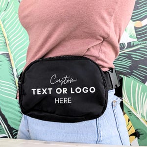 May include: Black fanny pack with the text "Custom TEXT OR LOGO HERE" in white. The bag has a zipper closure and an adjustable black strap with a plastic buckle. The person is wearing a pink shirt and blue jeans.
