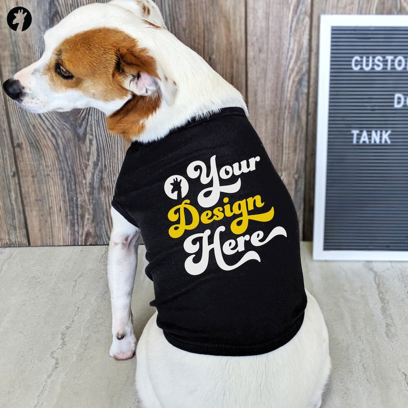 K&ouml;nnte beinhalten: Ein wei&szlig;er Hund tr&auml;gt ein schwarzes T-Shirt mit dem Text "Your Design Here" in Gelb. Der Hund sitzt auf einem Fliesenboden vor einer Holzwand.