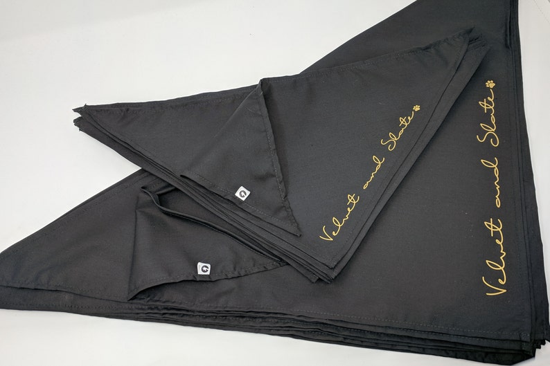 May include: A stack of black bandana dog bandanas with gold embroidery that reads "Velvet and Slate".