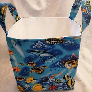 Pode incluir: Uma caixa de armazenamento de tecido azul com alças brancas. A caixa tem uma estampa colorida de peixes, golfinhos e recifes de coral.