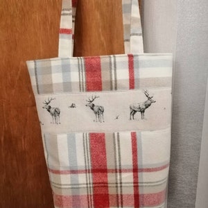 Puede incluir: Una bolsa de mano de cuadros rojos y blancos con un panel de tela beige que presenta una fila de tres siluetas de ciervos en blanco y negro. La bolsa tiene dos asas de cuadros rojos y blancos.