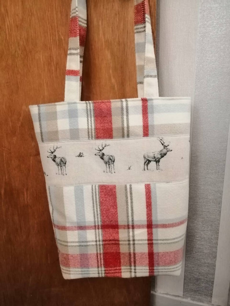 Puede incluir: Una bolsa de mano de cuadros rojos y blancos con un panel de tela beige que presenta una fila de tres siluetas de ciervos en blanco y negro. La bolsa tiene dos asas de cuadros rojos y blancos.