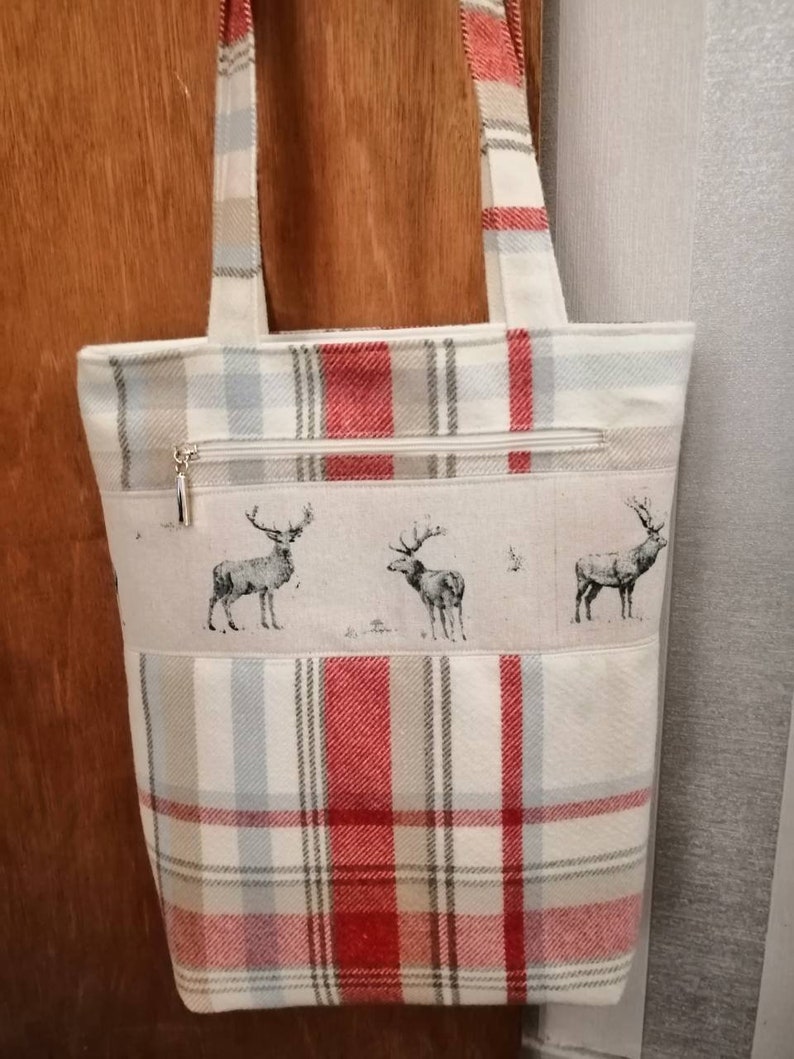 Puede incluir: Una bolsa de mano de cuadros rojos y blancos con un bolsillo con cremallera y un dise&ntilde;o de estampado de ciervos. La bolsa tiene dos asas.