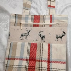 Puede incluir: Una bolsa de mano beige con tejido de cuadros rojos, marrones y azules. La bolsa tiene un bolsillo con cremallera y un dise&ntilde;o de ciervos en la parte delantera.