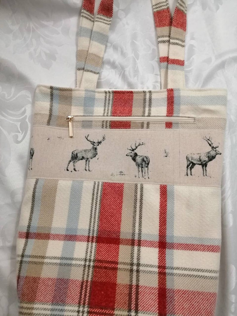 Puede incluir: Una bolsa de mano beige con tejido de cuadros rojos, marrones y azules. La bolsa tiene un bolsillo con cremallera y un dise&ntilde;o de ciervos en la parte delantera.
