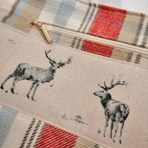 Puede incluir: Una bolsa de mano de lona a cuadros beige y rojo con cierre de cremallera y tirador de cremallera dorado. La bolsa presenta un dise&ntilde;o de dos ciervos negros y blancos sobre un panel de lona beige.