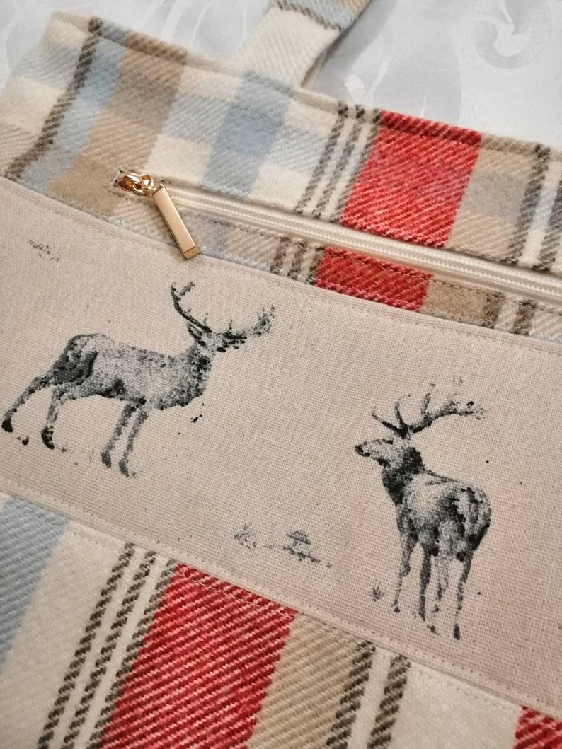 Puede incluir: Una bolsa de mano de lona a cuadros beige y rojo con cierre de cremallera y tirador de cremallera dorado. La bolsa presenta un dise&ntilde;o de dos ciervos negros y blancos sobre un panel de lona beige.