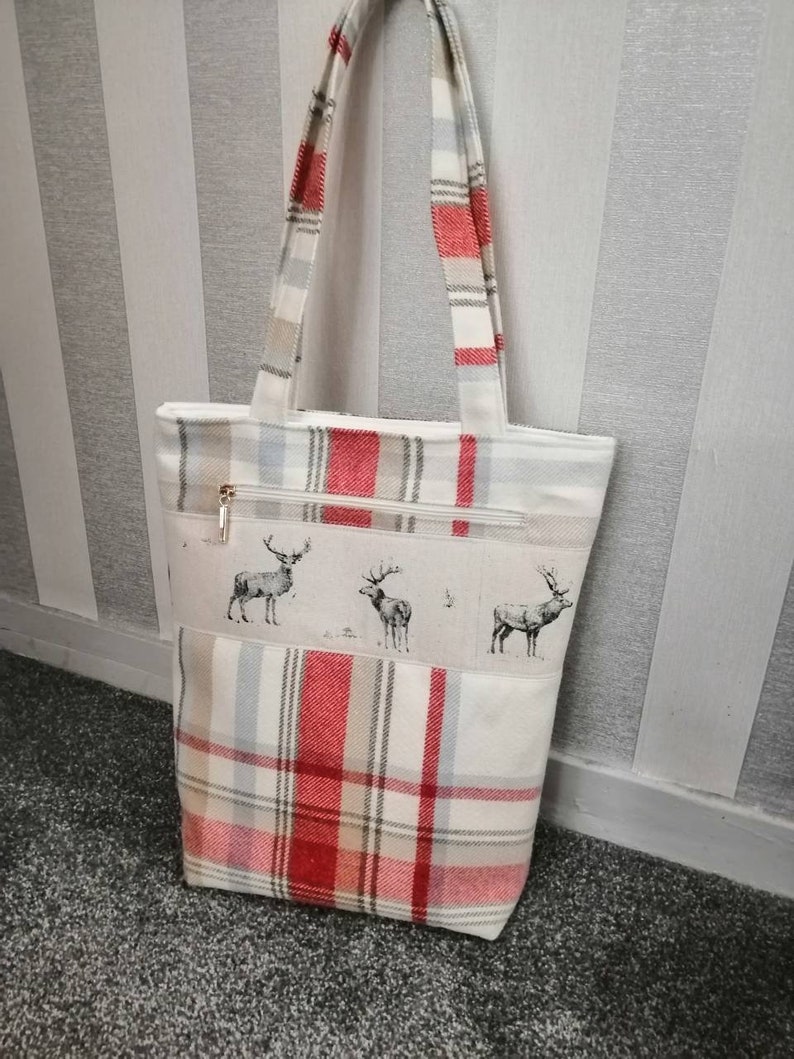 Puede incluir: Una bolsa de mano de cuadros rojos y blancos con cierre de cremallera y un dise&ntilde;o de estampado de ciervo. La bolsa tiene dos asas.