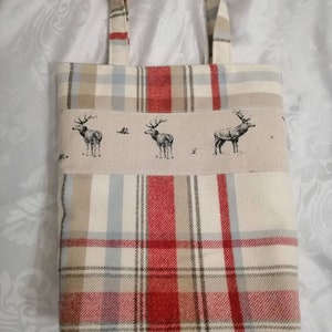 Puede incluir: Una bolsa de mano de cuadros rojos, blancos y marrones con un panel de tela beige que presenta un patr&oacute;n repetido de ciervos estilizados. La bolsa tiene dos asas de tela.