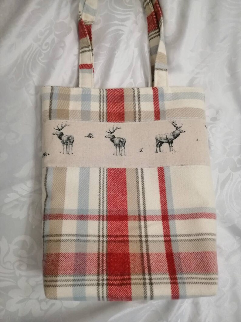 Puede incluir: Una bolsa de mano de cuadros rojos, blancos y marrones con un panel de tela beige que presenta un patr&oacute;n repetido de ciervos estilizados. La bolsa tiene dos asas de tela.