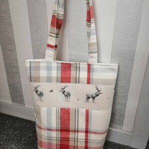 Puede incluir: Una bolsa de mano blanca con tela de cuadros rojos y beige. La bolsa tiene un panel de tela blanca con una fila de tres siluetas de ciervos negras y blancas. La bolsa tiene dos asas blancas.