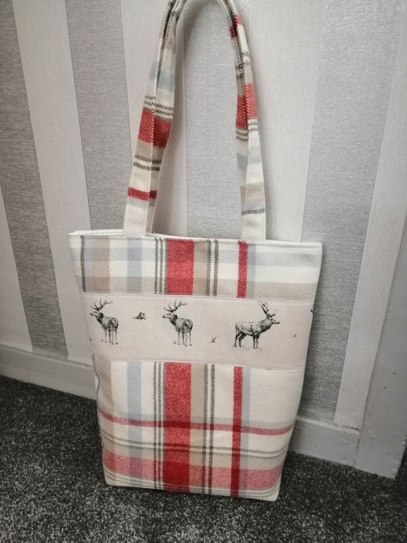 Puede incluir: Una bolsa de mano blanca con tela de cuadros rojos y beige. La bolsa tiene un panel de tela blanca con una fila de tres siluetas de ciervos negras y blancas. La bolsa tiene dos asas blancas.