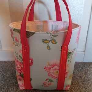 May include: A light blue tote bag with a floral print and red handles. The bag is lined with a pink and white checkered fabric.