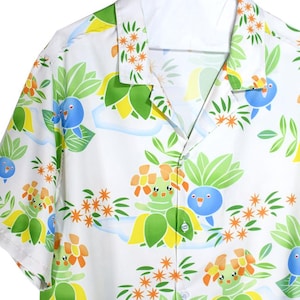 Peut inclure: Chemise blanche boutonnée avec un motif coloré de personnages de dessins animés, de fleurs et de feuilles. La chemise a des manches courtes et un col classique. La chemise porte le texte "POP Comet" en haut à droite.