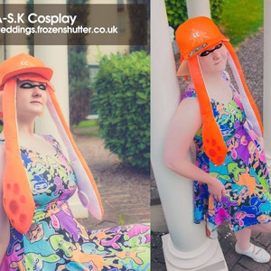May include: A woman wearing an orange and white squid hat and a colorful dress with a squid pattern. The dress has a variety of colors including blue, green, purple, and pink. The hat has a white brim and orange tentacles. The woman is standing in front of a white pillar.