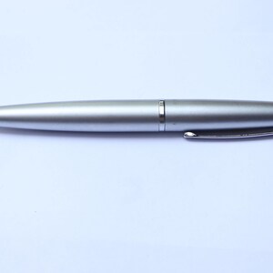Puede incluir: Un bolígrafo plateado con un acabado metálico liso. El bolígrafo tiene una punta, un clip plateado y un anillo en el centro. El bolígrafo está diseñado para escribir y está sobre un fondo blanco.