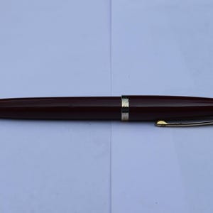 Puede incluir: Un bolígrafo burdeos con detalles dorados. El bolígrafo tiene un diseño elegante y aerodinámico con una punta y un clip dorados. El cuerpo del bolígrafo es de color burdeos oscuro y tiene una punta negra.