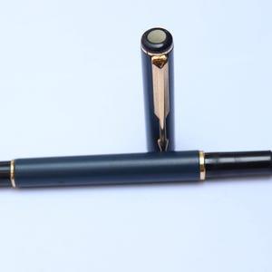 Könnte beinhalten: Dunkelblauer Füllfederhalter mit goldenen Akzenten und silberner Feder. Die Kappe ist abgenommen. Schreibgerät, ideal für Kalligraphie oder den täglichen Gebrauch. Länge ca. 14 cm.