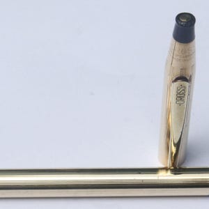 Könnte beinhalten: Ein goldfarbener Stift mit silberner Spitze und schwarzer Kappe. Die Kappe des Stifts steht aufrecht, und das Wort "CROSS" ist auf dem Stiftkörper sichtbar. Ein klassisches Schreibgerät.