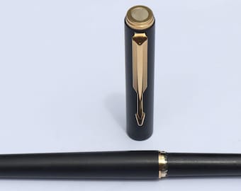 May include: A black fountain pen with gold-colored accents. The pen's cap is detached and stands upright, revealing a gold-colored clip. The pen's nib is visible, and the pen is lying on a white surface.