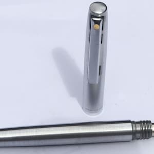 Puede incluir: Un bolígrafo plateado con un capuchón a juego. La punta del bolígrafo es visible y el capuchón está colocado junto al bolígrafo. El bolígrafo tiene una punta roja y un clip plateado. Es un instrumento de escritura.