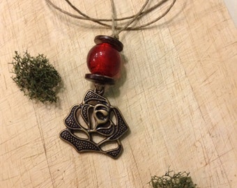 May include: A necklace with a bronze rose pendant and a red bead. The pendant is a detailed openwork design of a rose. The necklace has a brown cord and is displayed on a wooden surface with green moss.