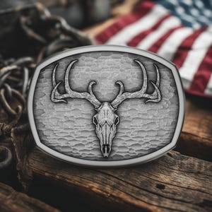 May include: Silver-toned oval belt buckle featuring a detailed deer skull with large antlers. The buckle has a textured background and a polished border. The background includes a blurred American flag and wooden surface.