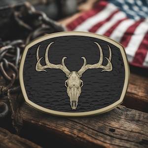 May include: An oval-shaped belt buckle with a detailed deer skull and antlers design. The skull is a light tan color, set against a textured black background, framed by a gold-colored border. The background includes a blurred American flag and wooden planks.