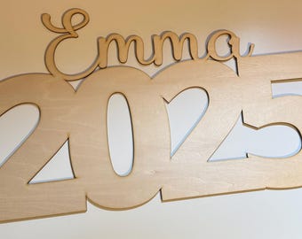 May include: Wooden sign with the name "Emma" and the year "2025" cut out. The sign is a light brown color.