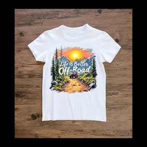 Könnte beinhalten: Weißes T-Shirt mit einem farbenfrohen Grafikdesign eines Sonnenuntergangs über einem Bergpfad und einem Fahrzeug. Der Text "Life is Better Off-Road" ist in Weiß und Blau dargestellt. Das Shirt besteht aus einem weichen Material.