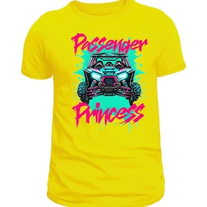 Könnte beinhalten: Ein gelbes T-Shirt mit einer Grafik eines Geländewagens und dem Text "Passenger Princess" in Pink und Türkis. Das Design umfasst eine detaillierte Illustration des Fahrzeugs mit großen Reifen und hellen Scheinwerfern.