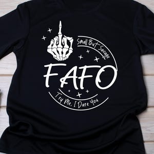 Puede incluir: Camiseta negra con un gráfico blanco que muestra una mano esquelética haciendo una peineta. El texto dice "FAFO" con "Small But Savage" y "Try Me, I Dare You" en un diseño circular.