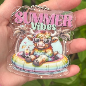 May include: A clear acrylic keychain with a colorful illustration. The design features a Highland cow wearing heart-shaped sunglasses and floating in a rainbow inner tube. The text "SUMMER Vibes" is displayed in pink and green.