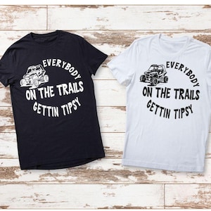 May include: Two short-sleeved t-shirts, one black and one white, with a graphic design. The design features an off-road vehicle and the text "EVERYBODY ON THE TRAILS GETTIN TIPSY" in a circular layout. The shirts are displayed on a wooden background.