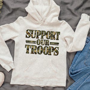 Könnte beinhalten: Ein hellgrauer Kapuzenpullover mit dem Text "SUPPORT OUR TROOPS" in einem Tarnmuster. Der Kapuzenpullover hat eine Vordertasche, eine Kapuze und lange Ärmel. Der Kapuzenpullover wird auf einer grauen Oberfläche mit einer Jeans präsentiert.