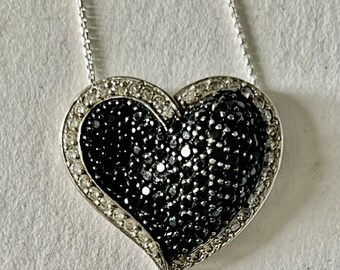 Puede incluir: Un collar de cadena de plata con un colgante en forma de corazón. El colgante está cubierto de pequeños diamantes negros y blancos.