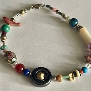 Puede incluir: Collar de cuentas multicolores con un colgante de botón grande negro y dorado. El collar presenta varias cuentas, incluyendo turquesa, rojo, blanco y azul.