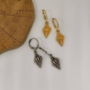 Peut inclure: Deux paires de boucles d'oreilles en forme de coquillage. Une paire est dorée avec des fermoirs à levier, et l'autre est argentée avec des fermoirs articulés. Les boucles d'oreilles sont présentées sur une surface blanche avec un fond en bois.