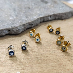 Könnte beinhalten: Eine Sammlung von Ohrsteckern mit Augenmotiv. Einige Ohrringe haben eine goldfarbene Fassung mit blau-weißem Auge, andere eine silberfarbene Fassung mit dunkelblauem Auge. Die Ohrringe sind klein und rund.