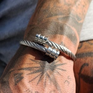 May include: Silver-toned cable bracelet with a screw-like clasp. The bracelet is worn on a wrist with intricate tattoos. The design features a twisted cable pattern and a secure closure.