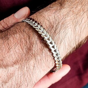 May include: Silver-toned bracelet with a wheat or leaf-like design. The bracelet is worn on a wrist, and the design is made up of repeating, connected elements. The bracelet is a bright silver color and appears to be made of metal.