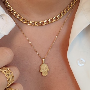Könnte beinhalten: Eine goldene Kettenkette mit einem Hamsa-Hand-Anhänger mit einem Davidstern. Die Kette ist über eine dickere goldene Kette gelegt. Die Person trägt ein weißes Hemd und einen Ring mit einem türkisfarbenen Stein.