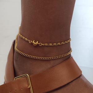 Può includere: Cavigliera dorata con due catenine sottili. La catena superiore ha un ciondolo a forma di luna crescente. Il gioiello è indossato con un sandalo in pelle marrone. Un accessorio alla moda.