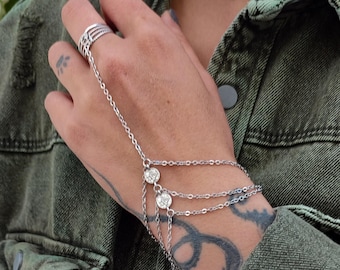 May include: A silver hand chain bracelet with a ring and three strands of chain. The bracelet features two round, clear gemstones and is connected to a ring with a multi-band design. The jewelry is worn on a hand.