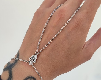 May include: Silver hand chain bracelet featuring a Hamsa hand charm. The bracelet connects to a finger ring with delicate chain links. The Hamsa hand charm is detailed with black accents.