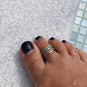 May include: Close-up of a foot with black nail polish and gold-colored toe rings with a turquoise band. The rings are stacked on the toe. The foot is wet, and the background is a light gray surface.