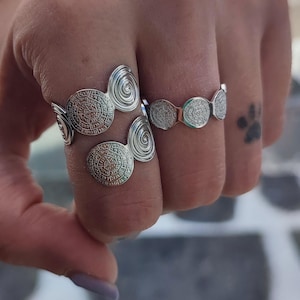 May include: Silver rings with detailed circular patterns are shown on a hand. The rings have spiral accents and are worn on several fingers. The rings' design is highlighted, showcasing their style. These rings are a fashion accessory.