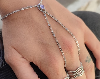 May include: Silver hand chain bracelet with a small purple gemstone and chain details. The hand is also adorned with two silver rings, one with a looped design and the other with a wavy pattern. The hand is resting on a dark gray surface.